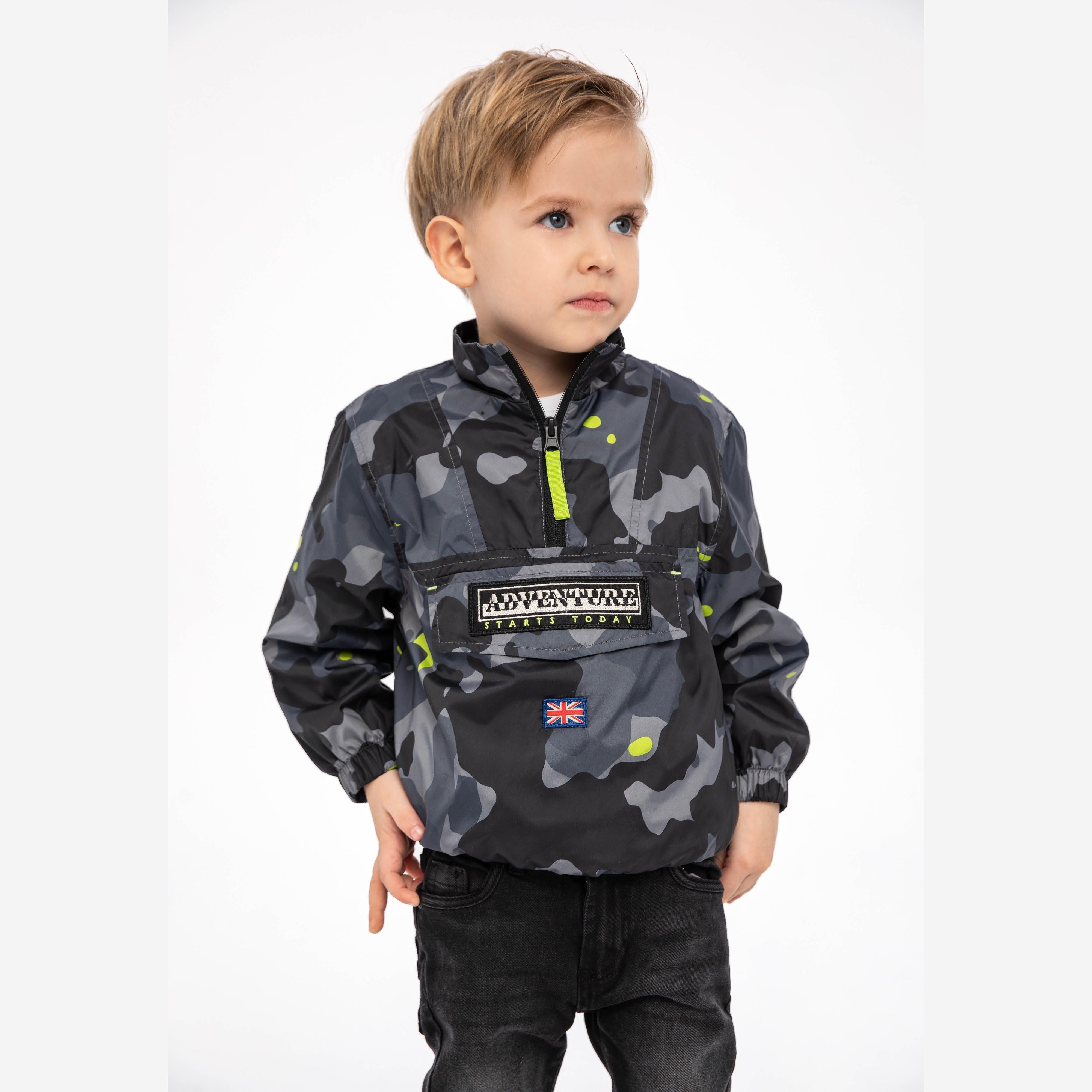 Young blonde boy wearing black camouflage windbreaker jacket with neon yellow half-zip, standing against white background