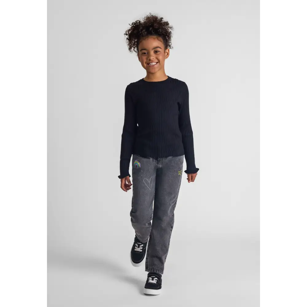 Young girl wearing black embroidered jeans with rainbow and star details, paired with navy ribbed sweater, walking against white studio background