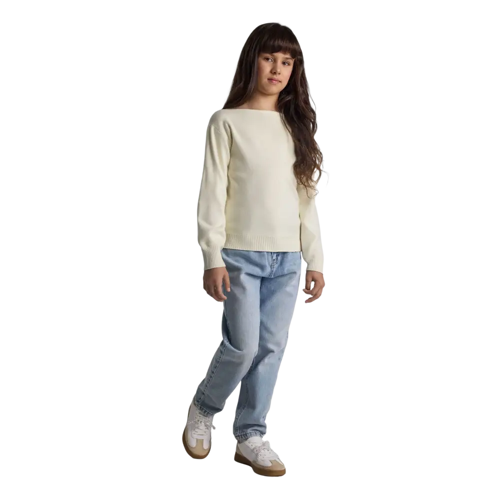 Young girl wearing cream textured knit jumper with boat neckline and light blue jeans, standing against black background with long brown hair