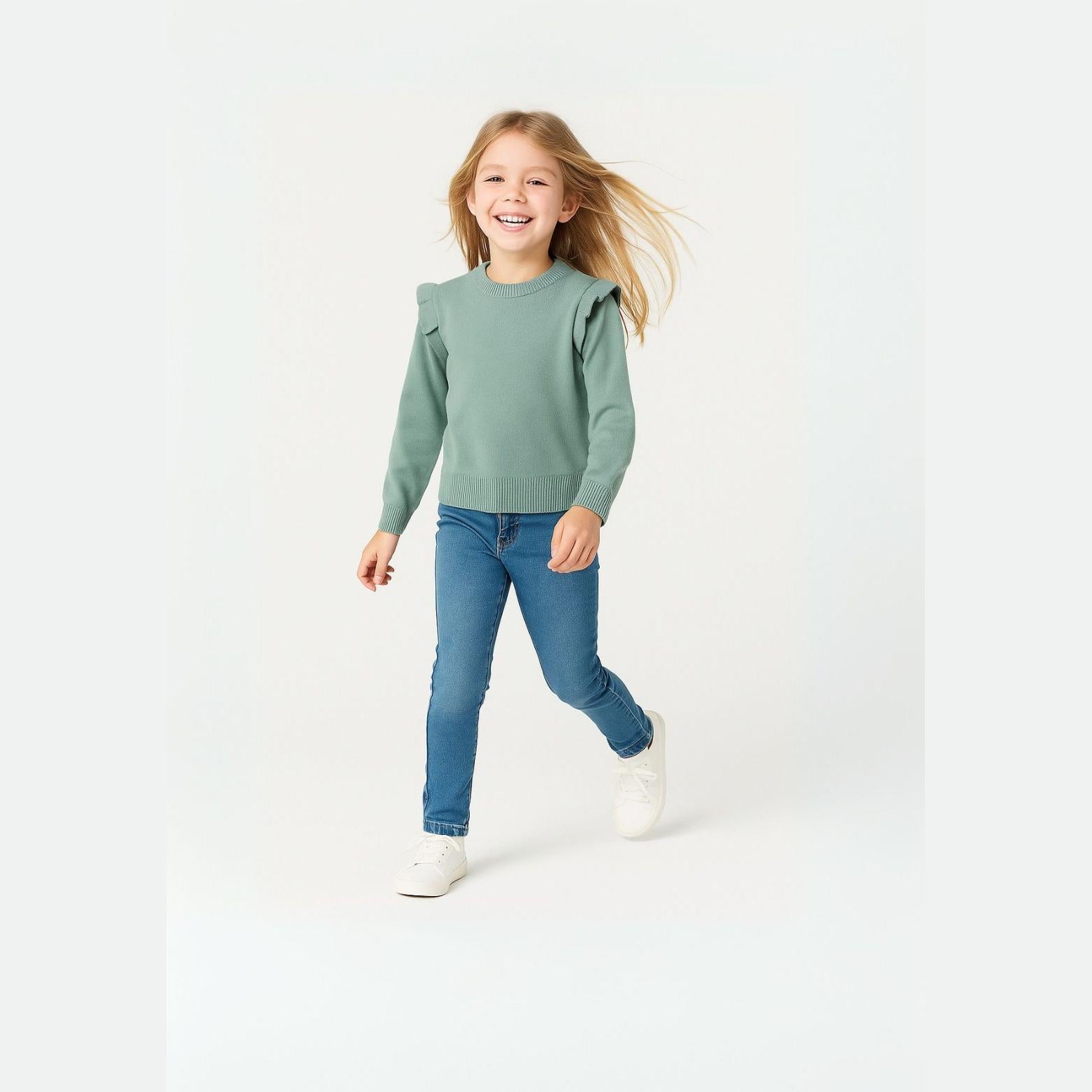 Young blonde girl in motion wearing mint green knitted jumper with shoulder frill details and blue jeans, white sneakers against light background