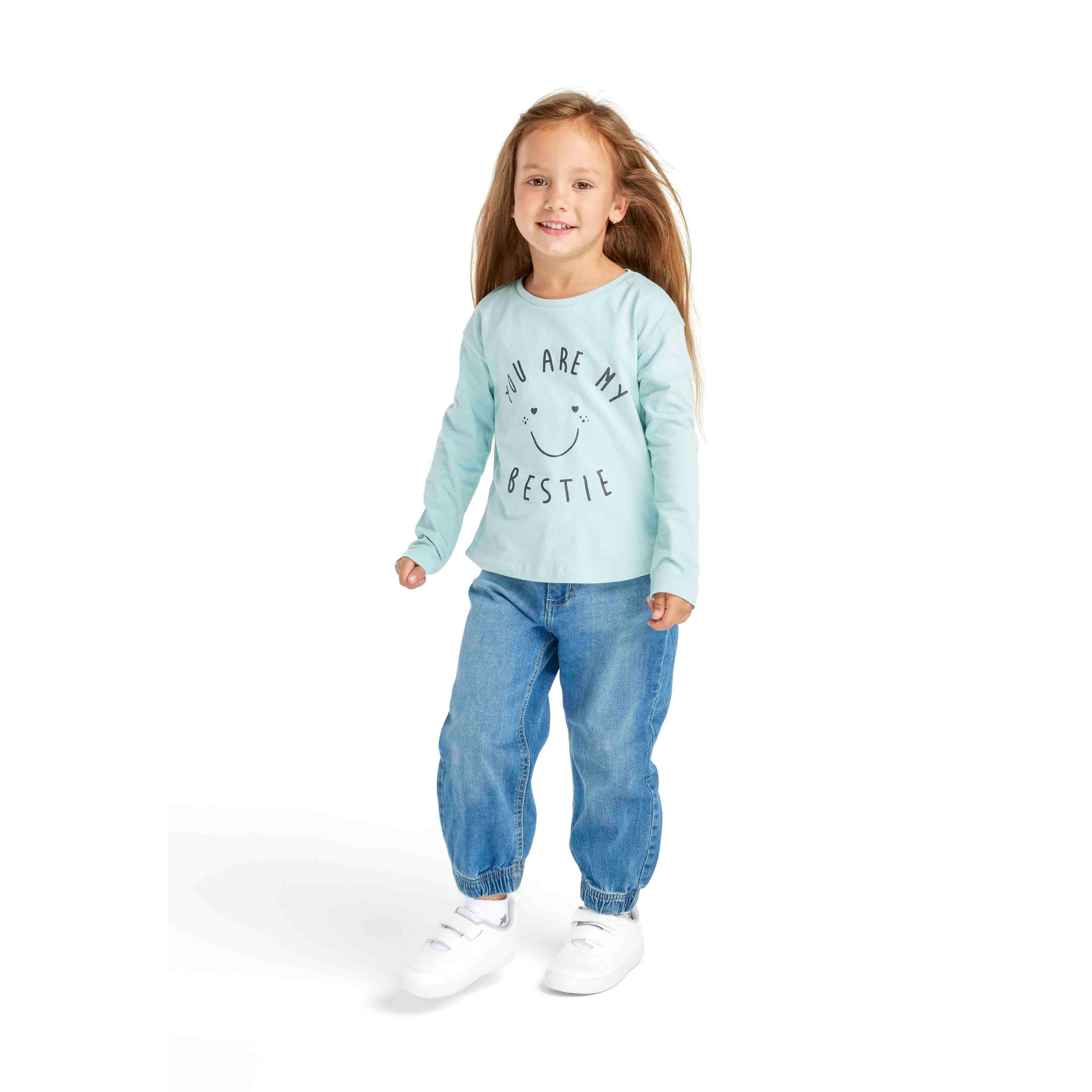 Young girl wearing mint long sleeve t-shirt with bestie smiley print and blue jeans, white sneakers, standing on white background