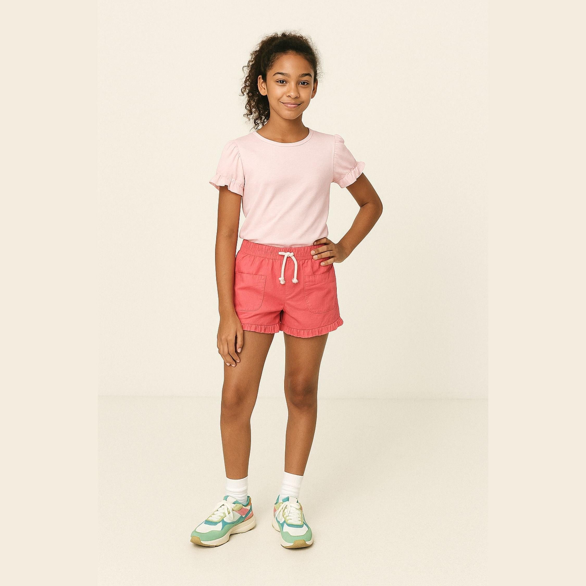 Young girl wearing pink poplin shorts with elasticated waistband, paired with light pink t-shirt and colorful sneakers, standing against cream background