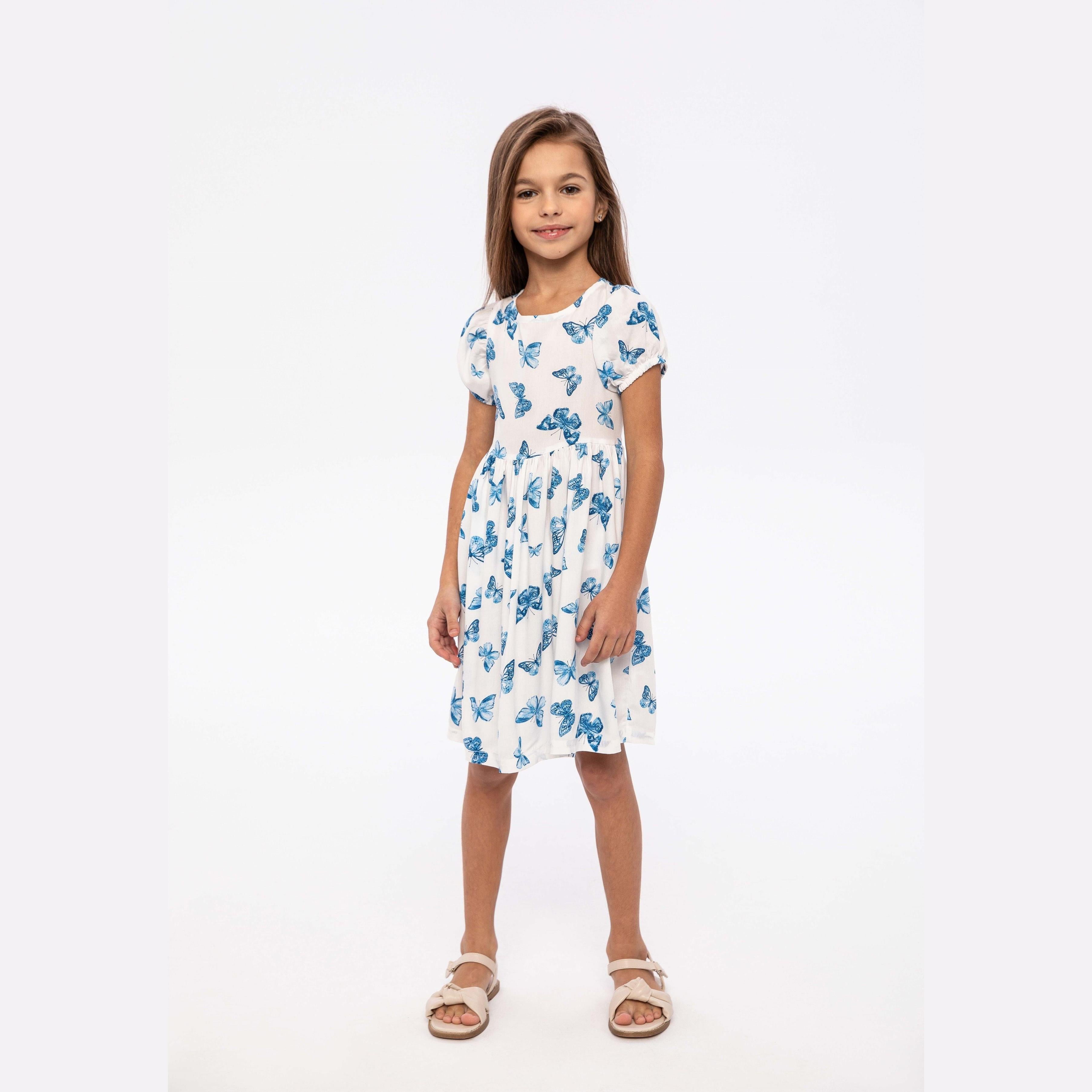 Young girl wearing white butterfly print dress with short sleeves, posing against white studio background with hands on hips