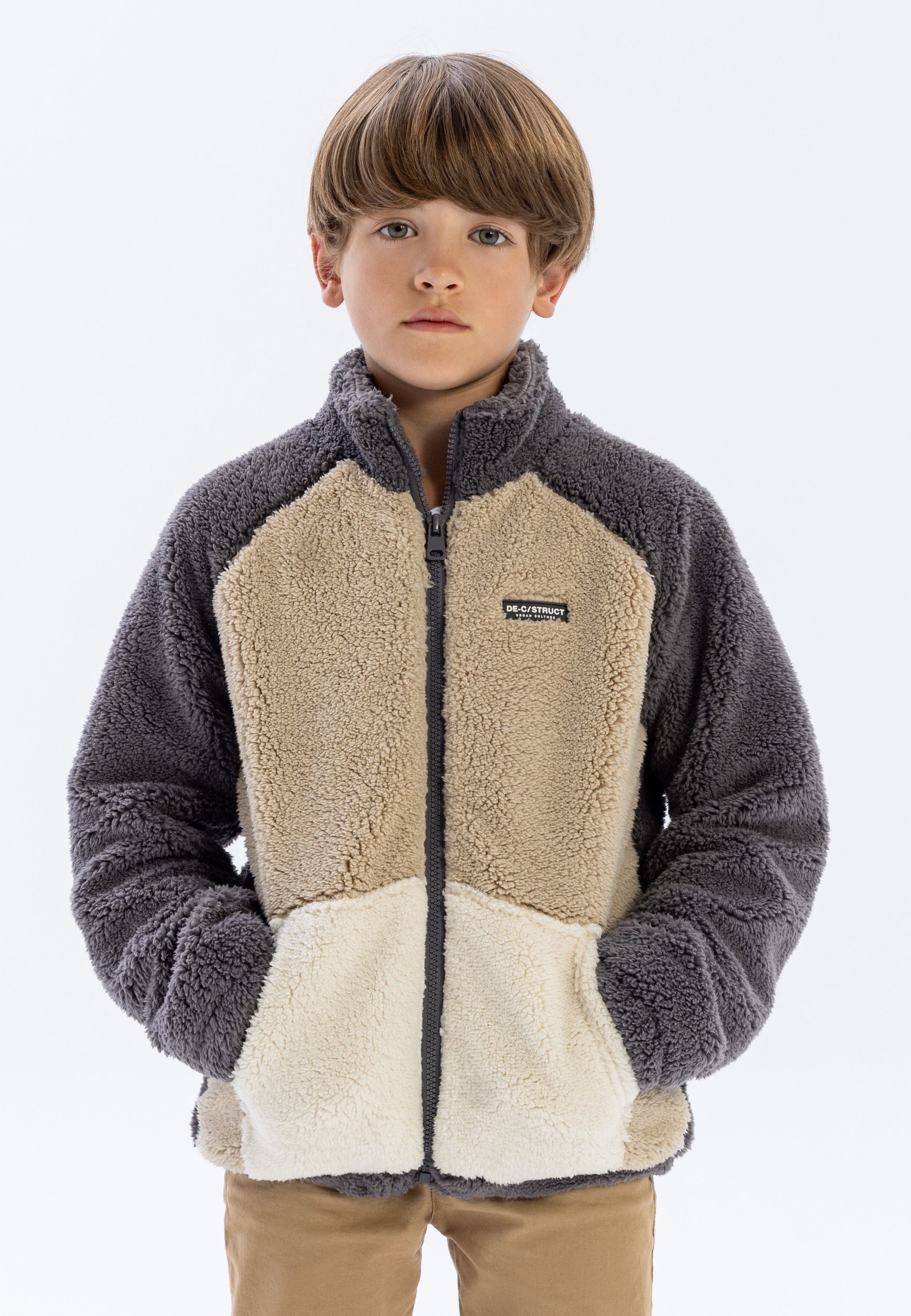 Boy wearing colourblock sherpa fleece jacket in brown, cream and natural tones with full zip and high collar, standing against white background